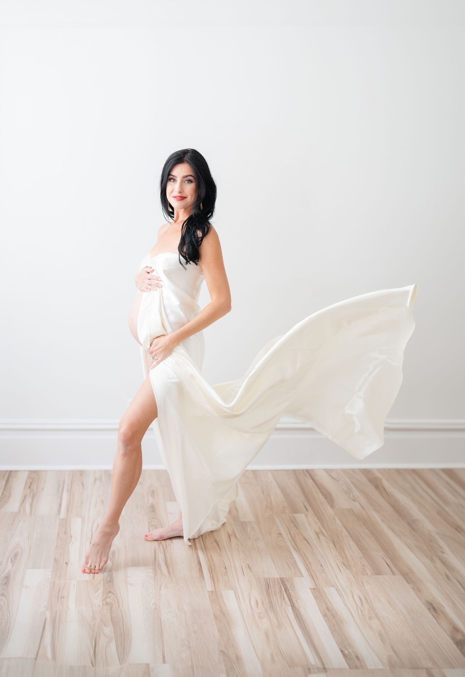 Expectant mother in flowing beige maxi dress poses gracefully against white background during maternity photoshoot.