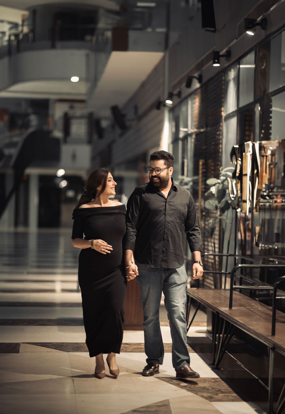 A couple walks together holding hands in a modern shopping mall hallway with retail displays.