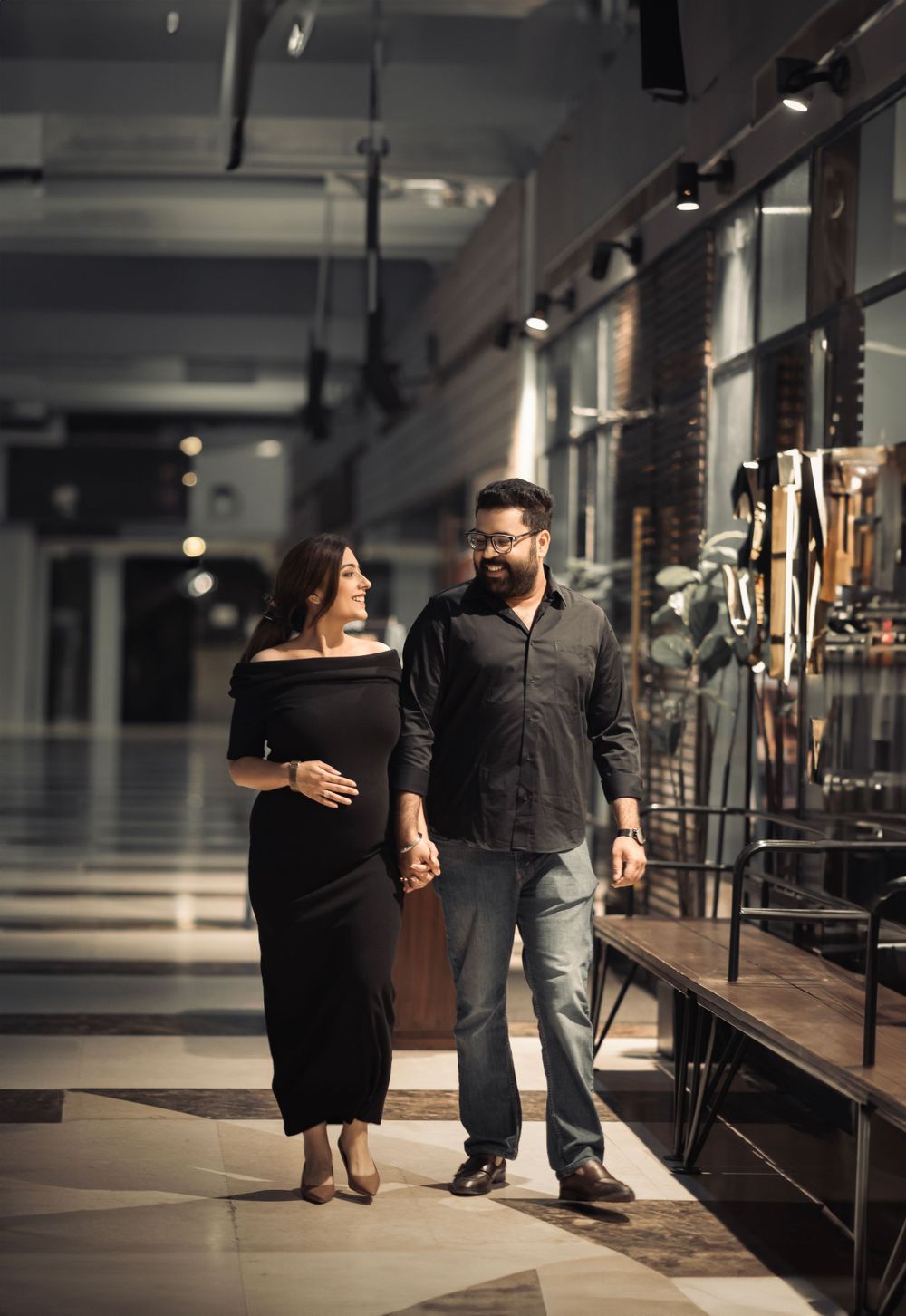 A couple strolls together in a mall interior, with the woman wearing a black dress.