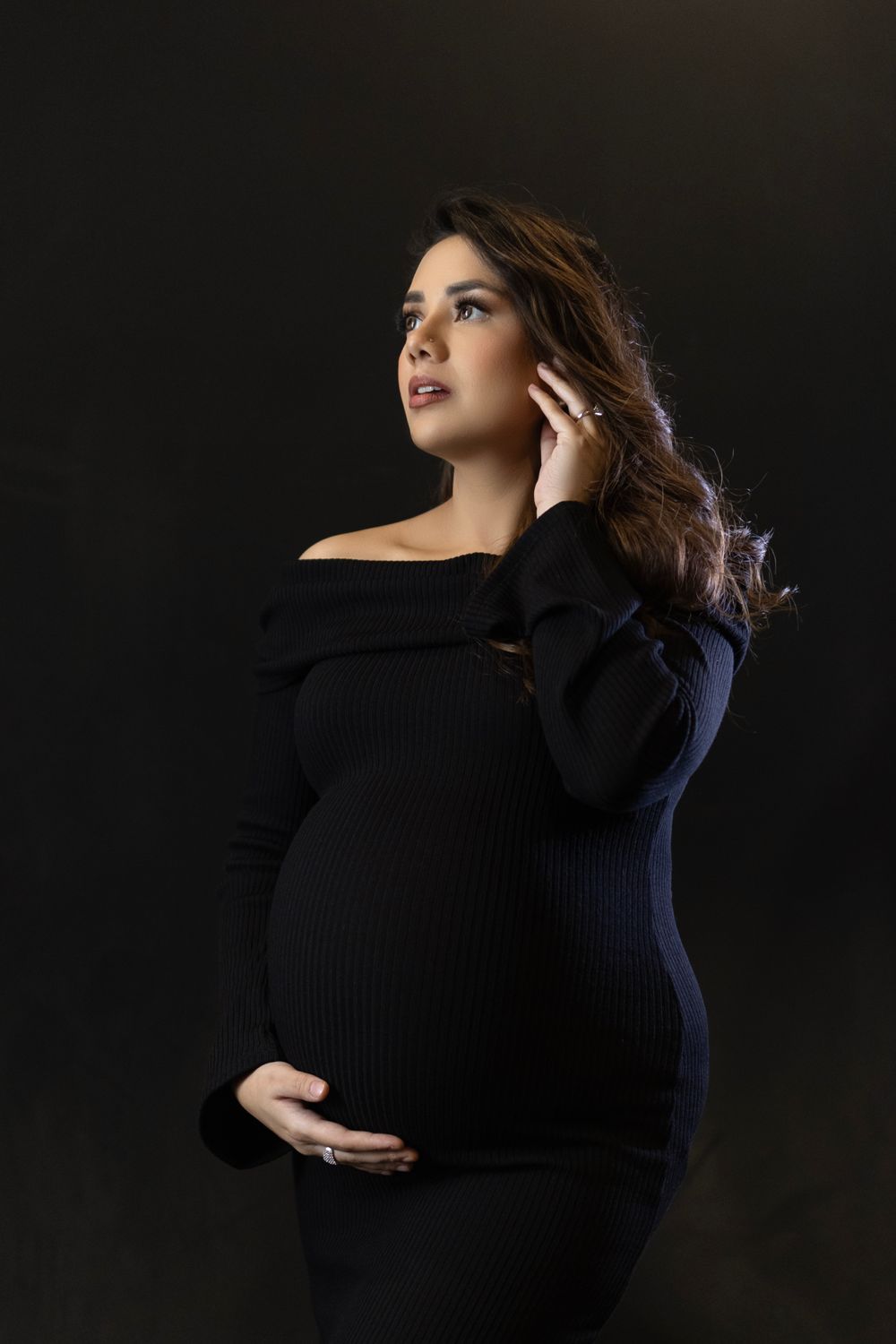 Dramatic black and white maternity portrait featuring an off-shoulder gown against dark background.
