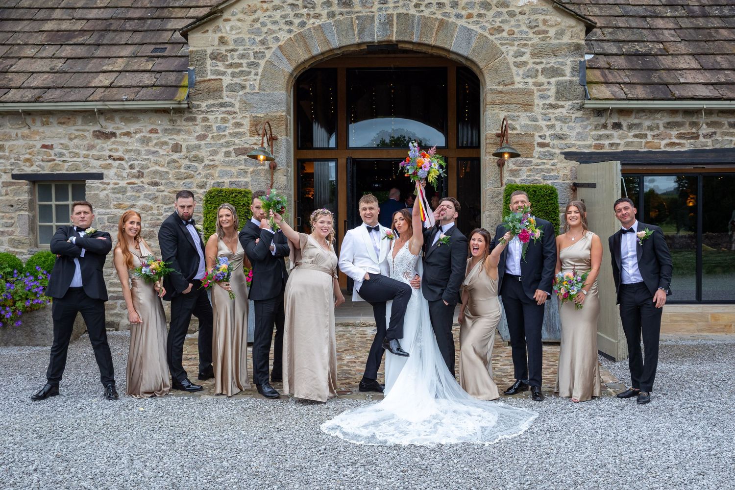 Wedding party poses outside stone venue, bridesmaids in gold dresses with groomsmen in black.