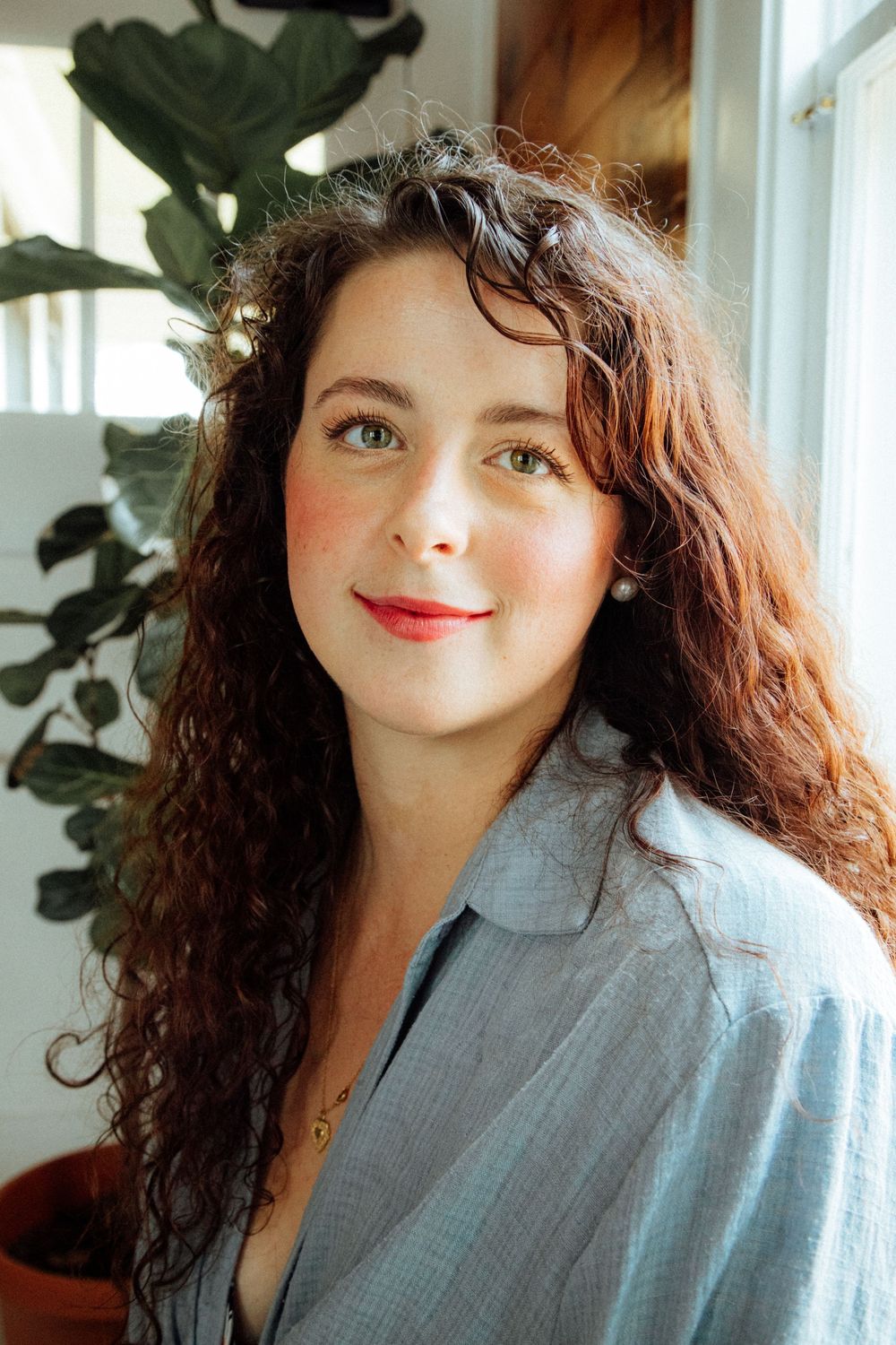 A person with long curly dark hair and red lipstick wearing a light blue button-up shirt poses near a window and houseplant.