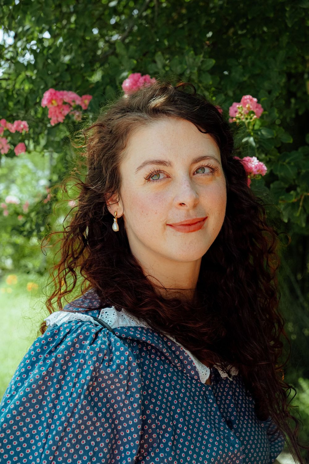 A young person in a blue polka dot dress poses outdoors with pink roses blooming in the background.