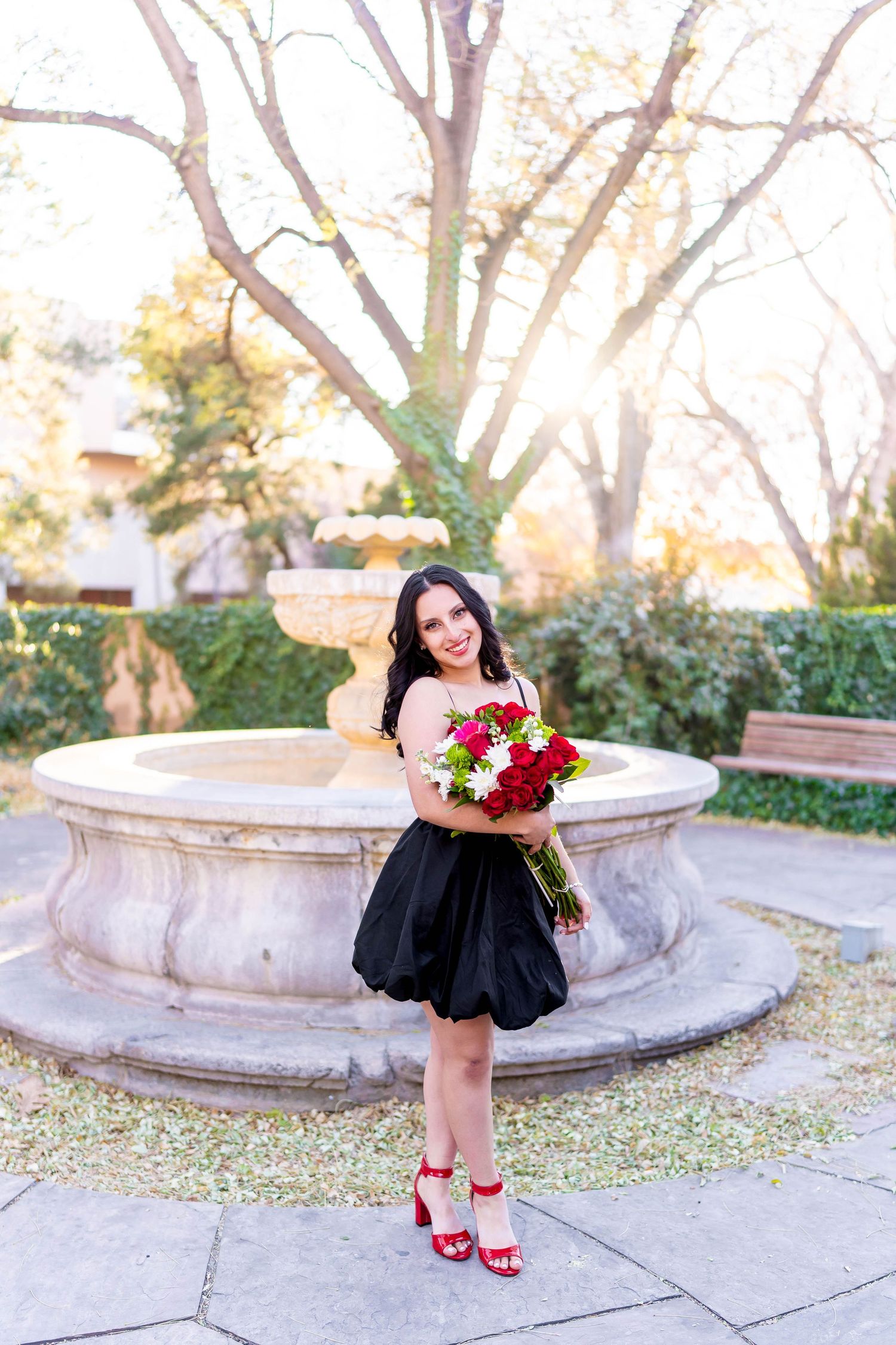 Elegant portrait of someone in a black dress holding red roses by a stone fountain.