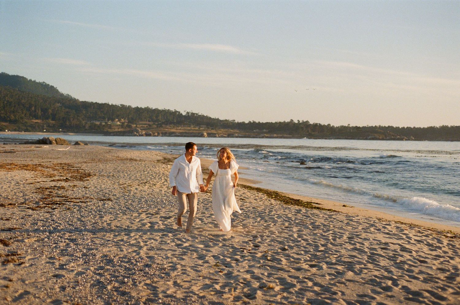 Couple in white outfits holding hands and walking along a sandy beach at sunset with ocean waves.