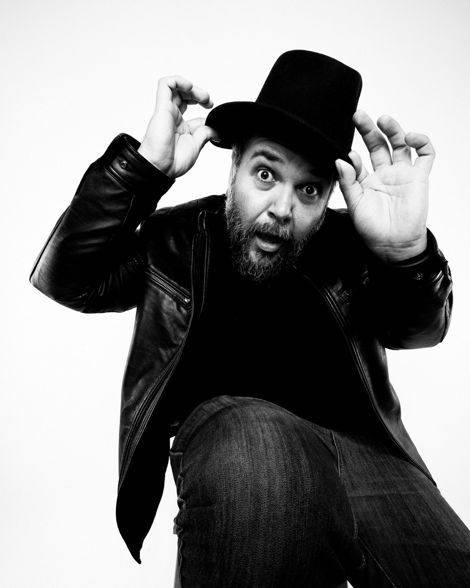 Black and white portrait photo of a playful man in a top hat and leather jacket making an expressive face against white background.