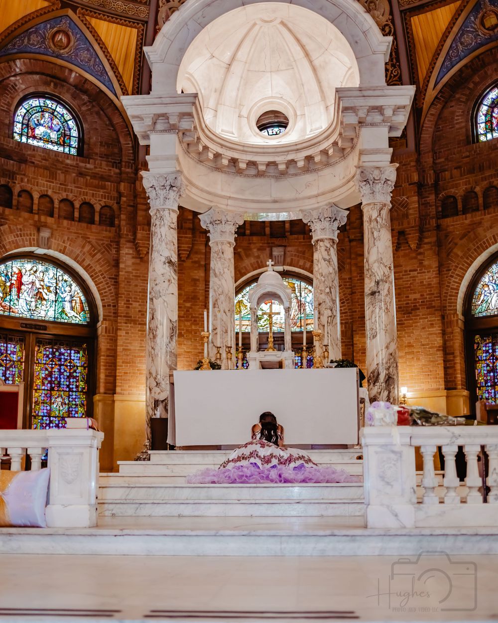 Ornate church interior with stained glass windows, marble columns and purple floral decorations at the altar.