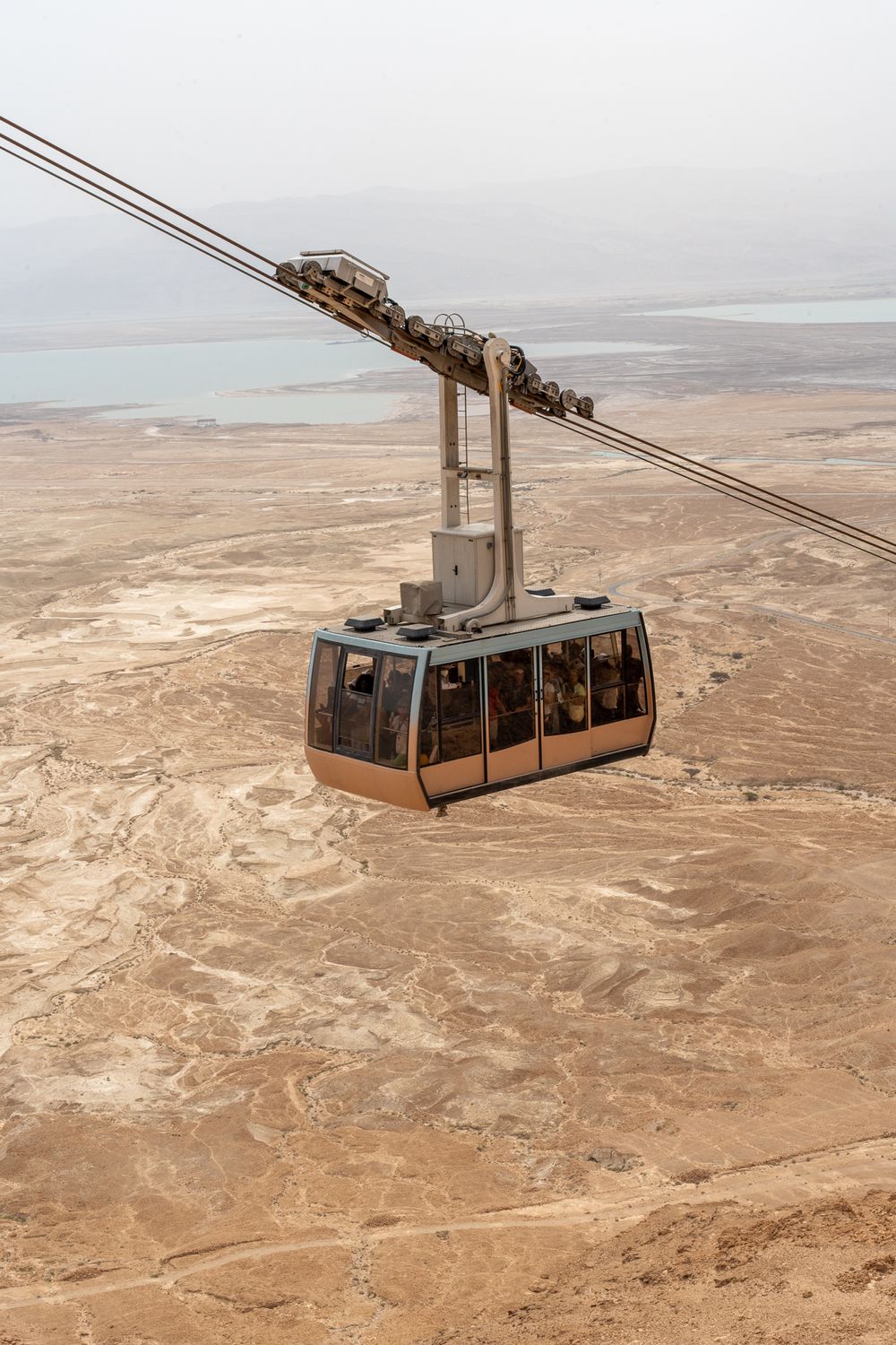 Cable car ascending over barren desert landscape with Dead Sea in background.
