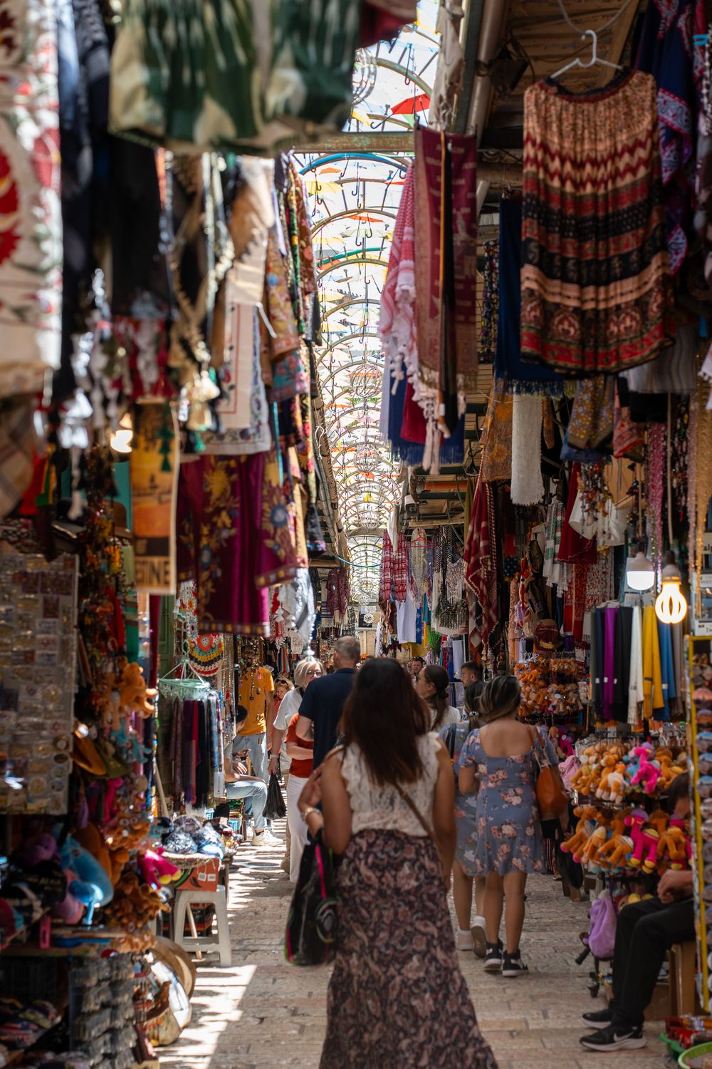 Bustling covered market bazaar with colorful textiles and merchandise lining narrow passageway.