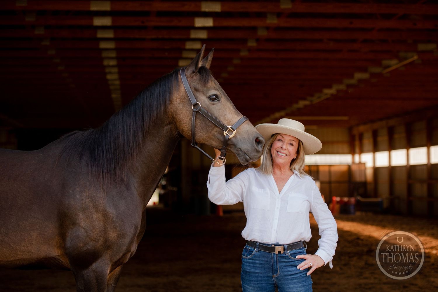 A dark horse and rider in western attire share a moment in a sunlit indoor arena.