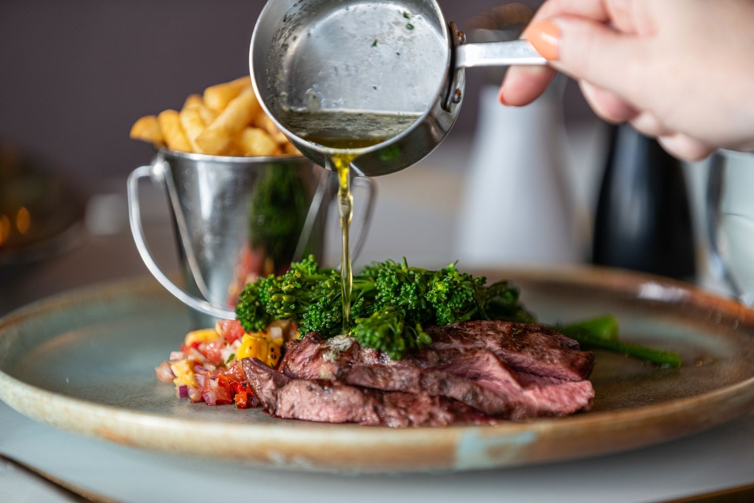 Sauce being poured over medium-rare steak with sides of French fries and garnish on ceramic plate.