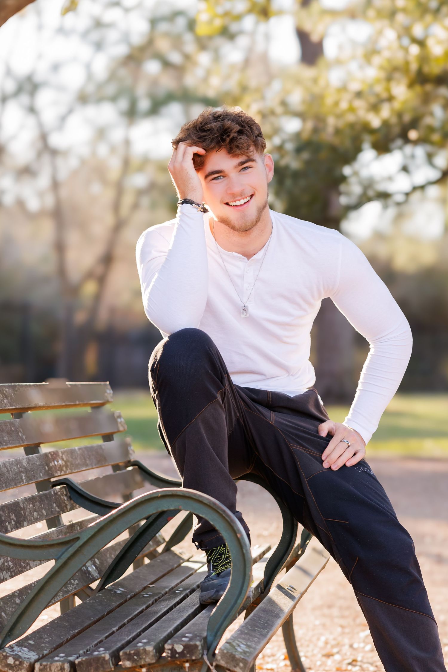 A person relaxes casually on a wooden park bench wearing a white long-sleeve shirt and dark pants.