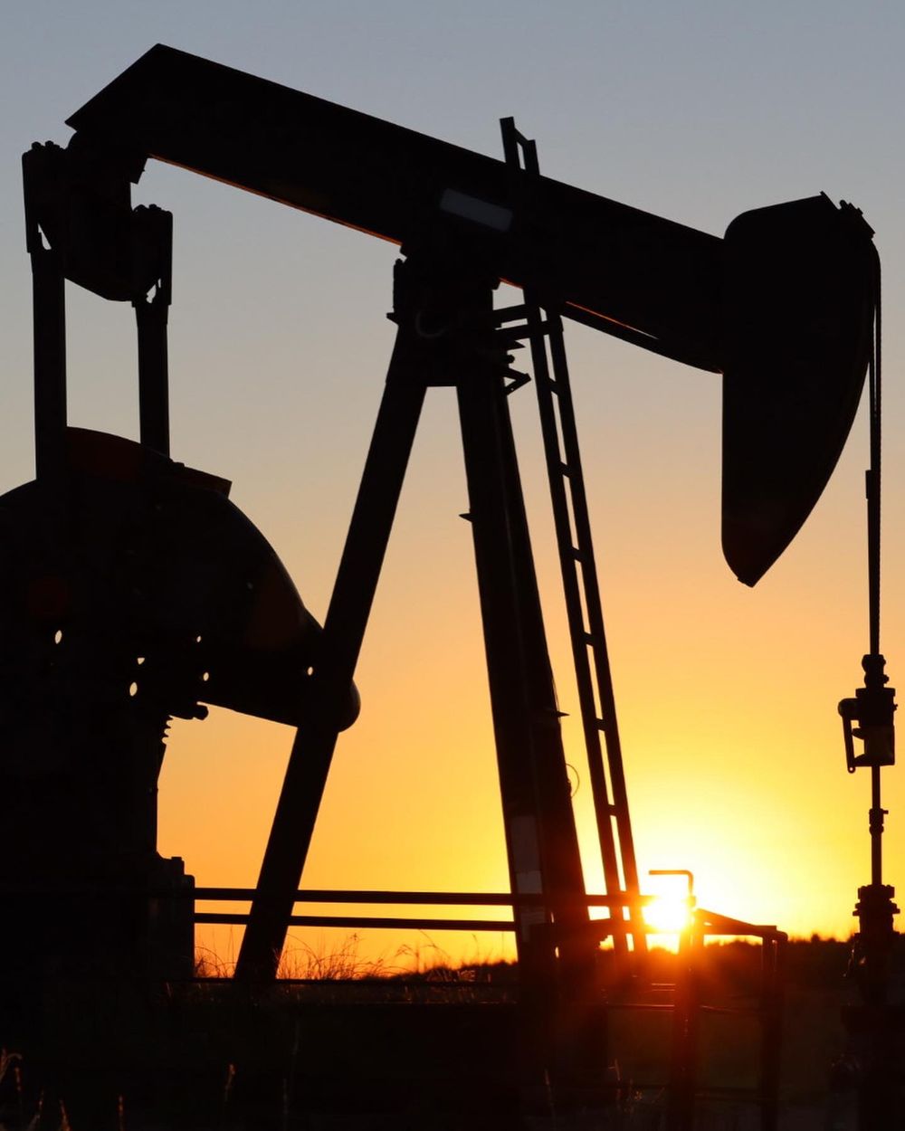Oil pump jack silhouetted against dramatic orange sunset sky.