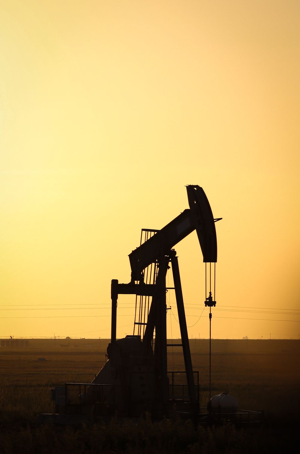 Oil pump jack silhouetted against warm yellow sunset sky in oil field.