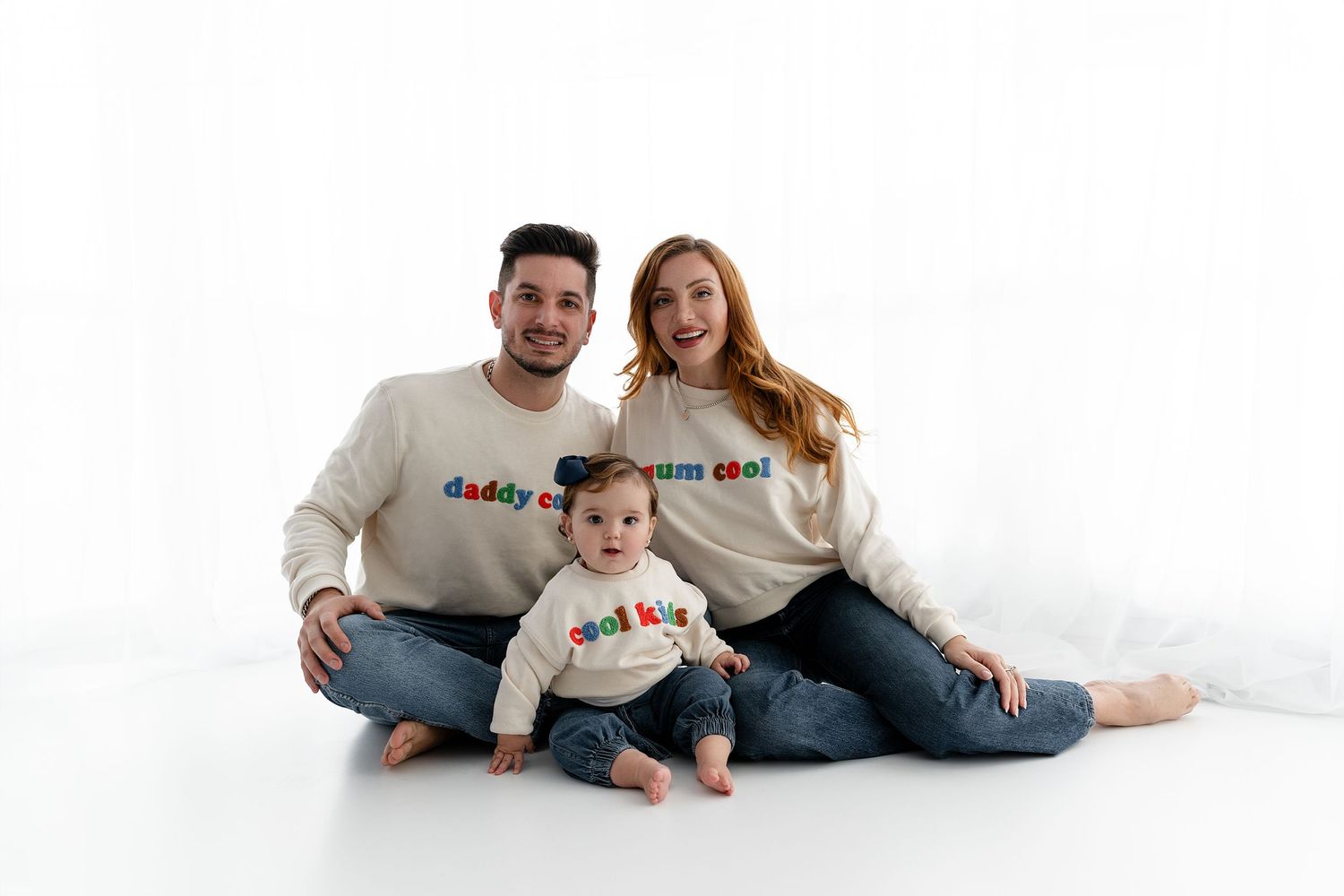 Family of three wearing matching sweaters sits together on white background.