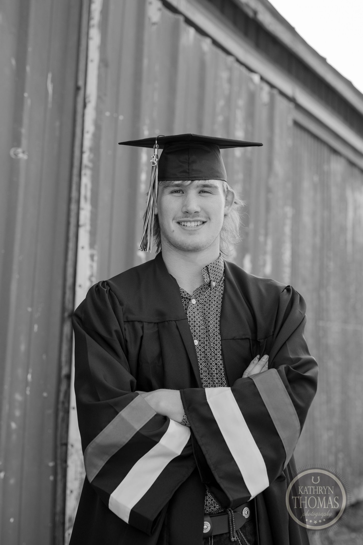Black and white portrait of graduate in cap and gown smiling against barn backdrop.
