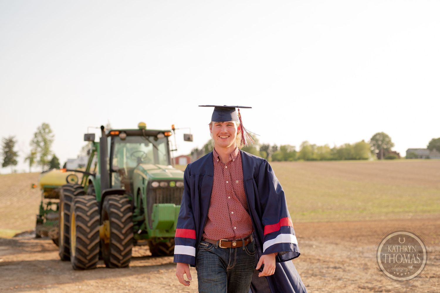 Graduate in academic regalia poses in front of tractor in sunlit field.