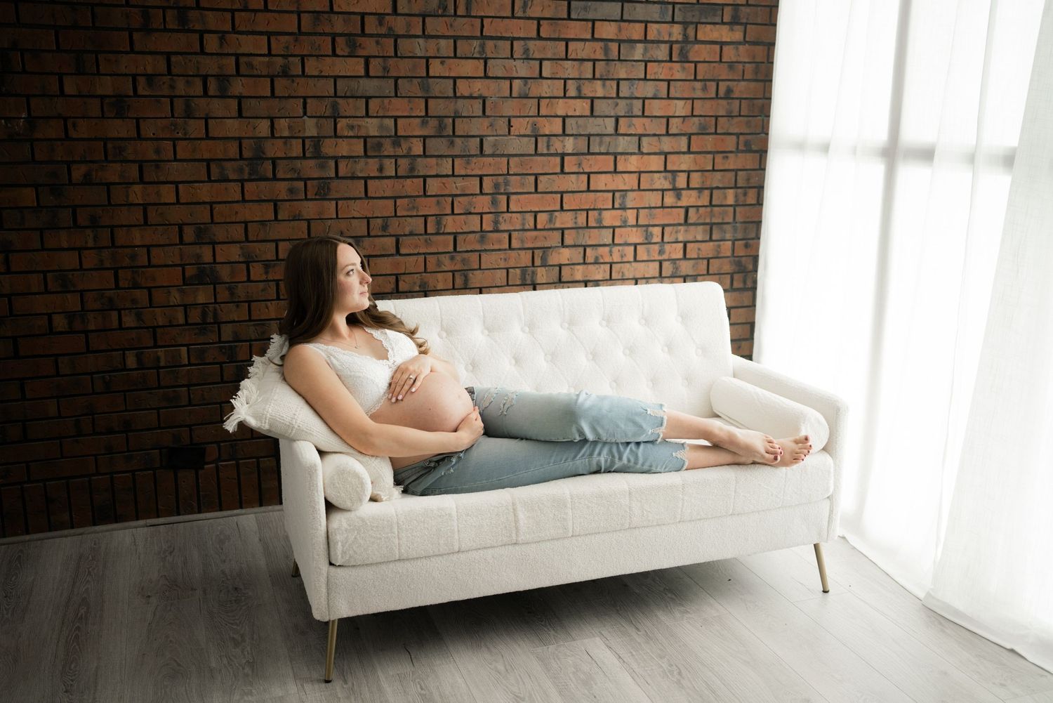 A relaxing maternity photoshoot featuring a white tufted couch against an exposed brick wall in natural light.