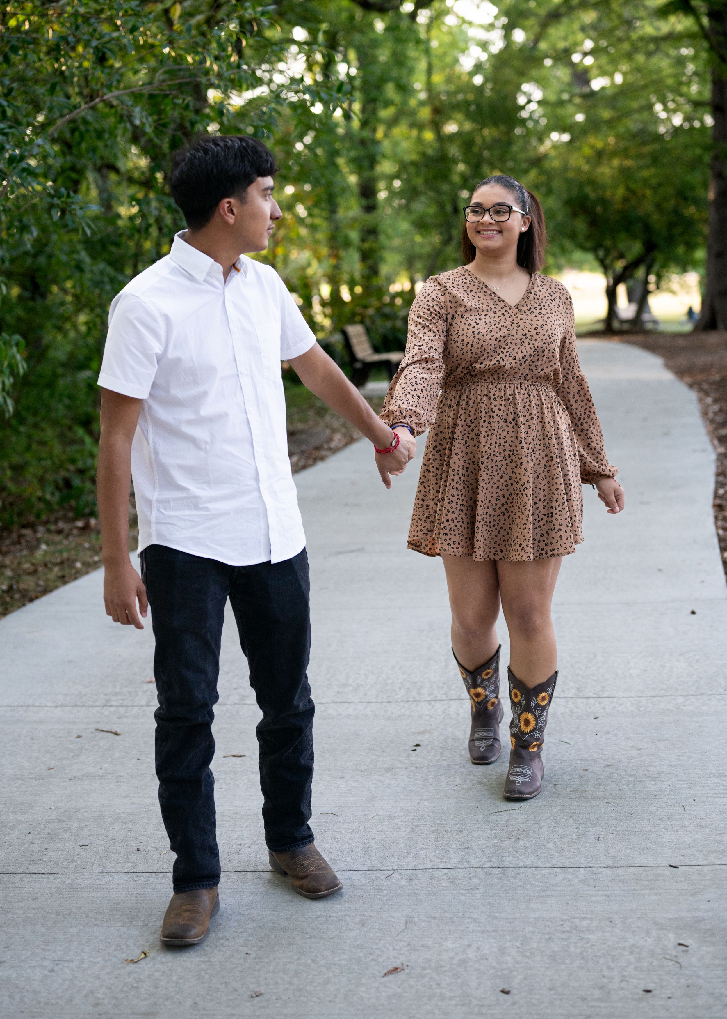 Couple holding hands while walking on a tree-lined path wearing casual outfits and cowboy boots.