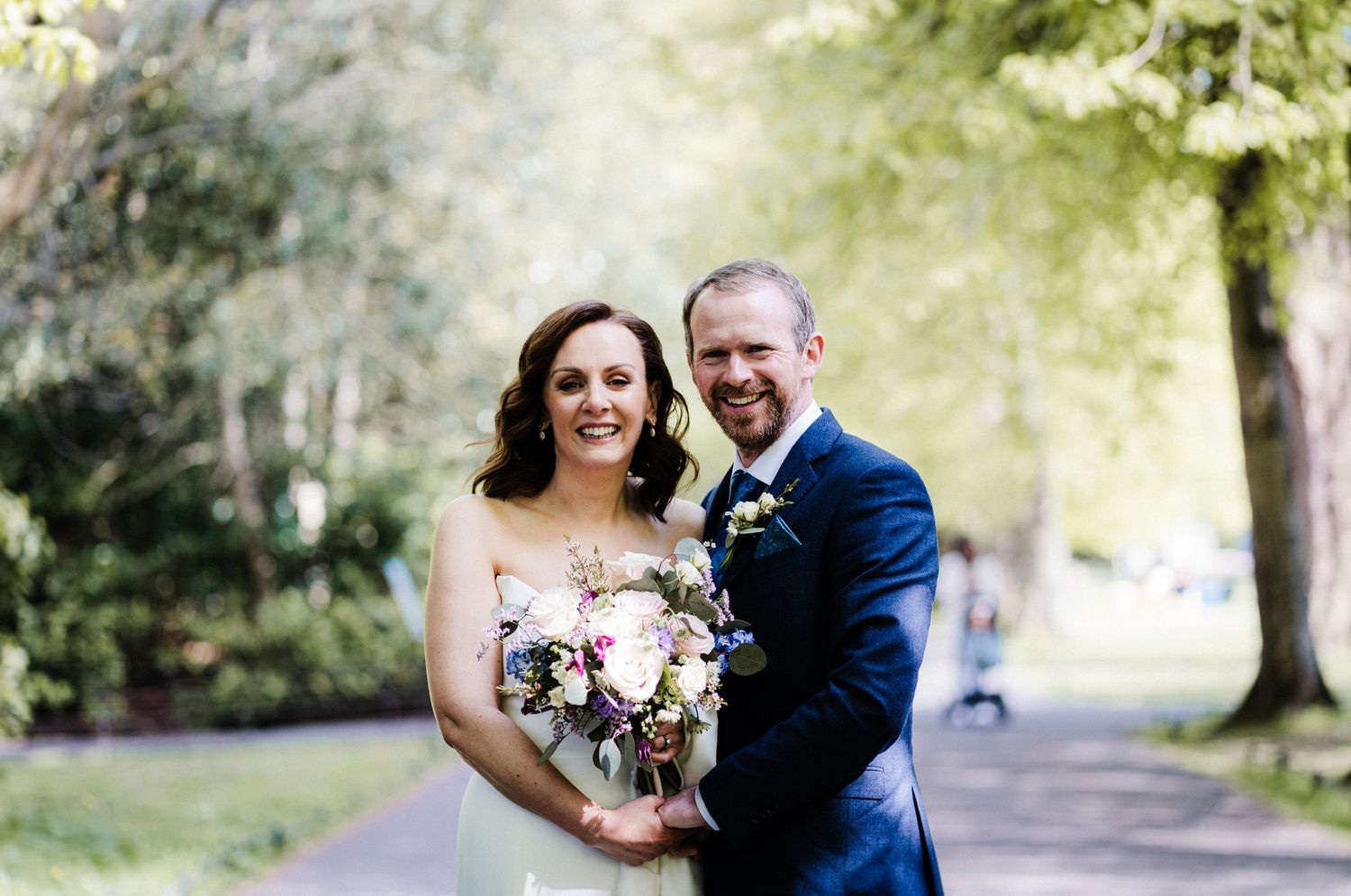 Happy couple poses outdoors in a park setting with a bridal bouquet and classic wedding attire.