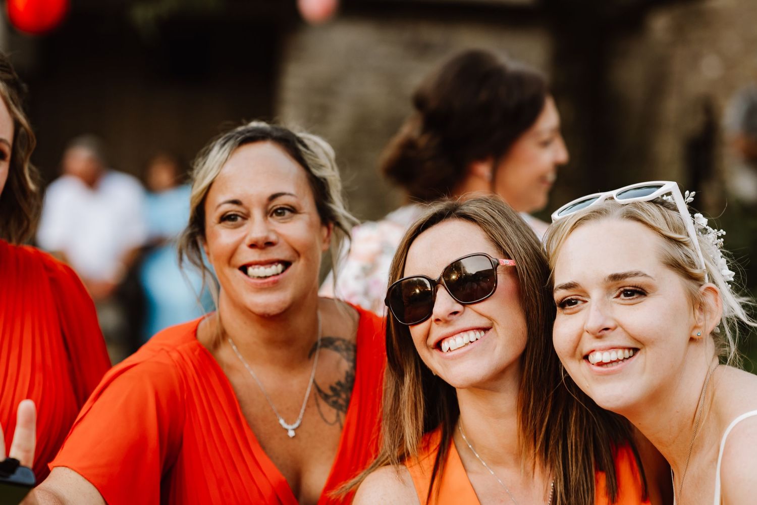 Three smiling women in orange outfits pose together outdoors at a sunny social event.