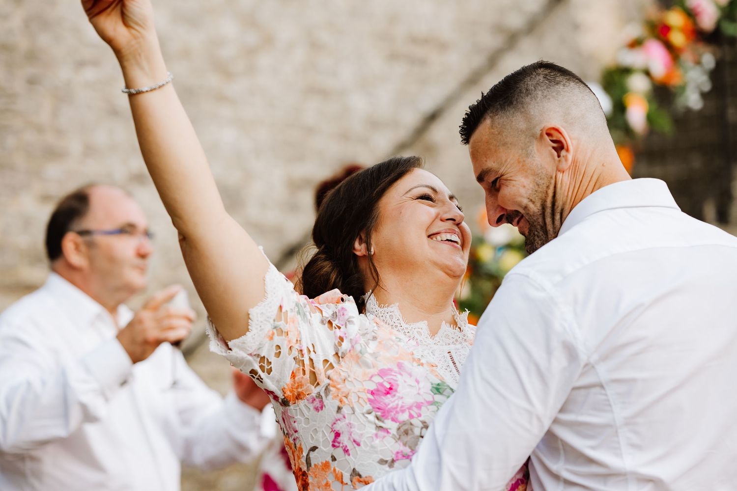 Joyful bride in floral dress raises hand while dancing with groom at outdoor wedding reception.