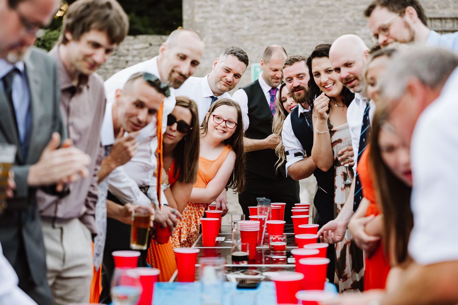 Wedding guests gather around beer pong table playing lawn games at outdoor reception party.
