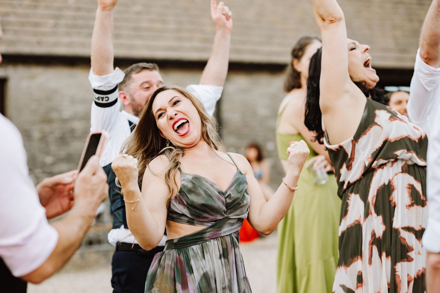 Excited wedding guests dance and celebrate with arms raised at lively outdoor reception party.