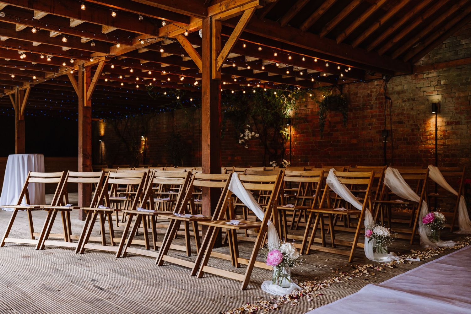 Rustic barn wedding ceremony setup with wooden folding chairs and warm string lights overhead at Elmhay, Orchardleigh estate, Frome