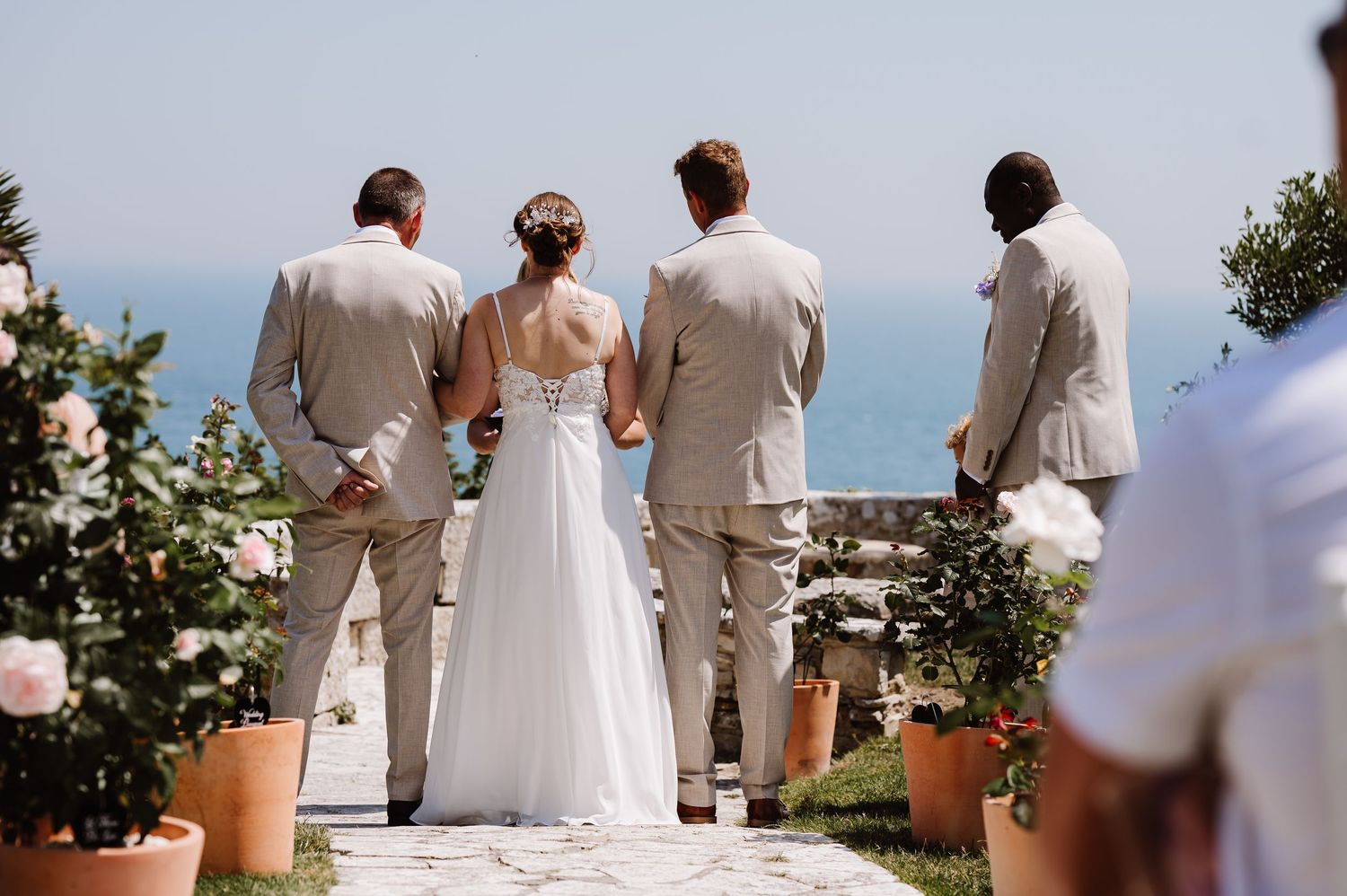 Bride and three groomsmen in beige suits standing overlooking the ocean at a seaside wedding. Pennsylvania Castle, Portland Dorset