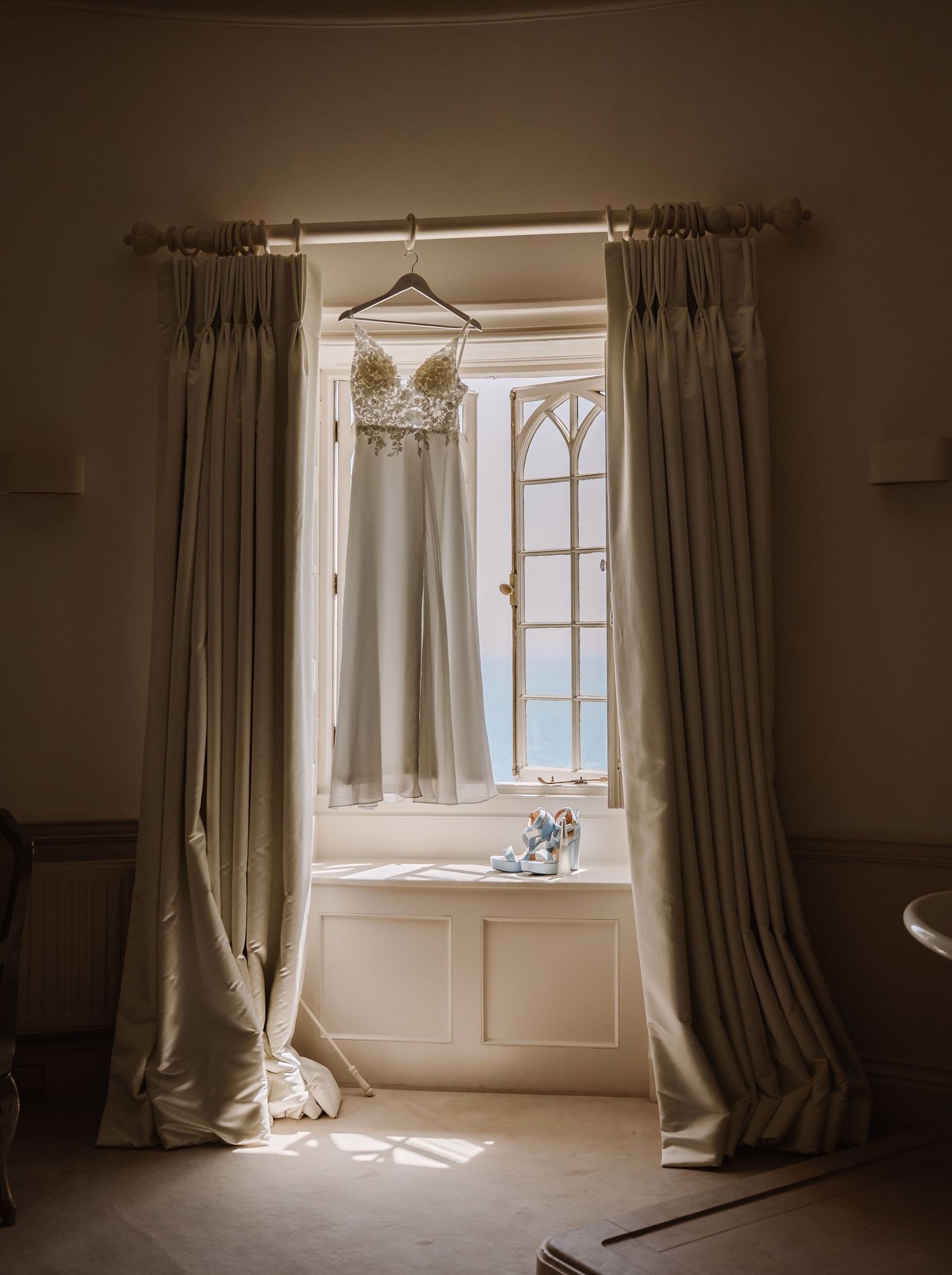 Pennsylvania Castle Portland Elegant wedding dress hanging in a sunlit Gothic window with blue heels on the sill at Pennsylvania Castle