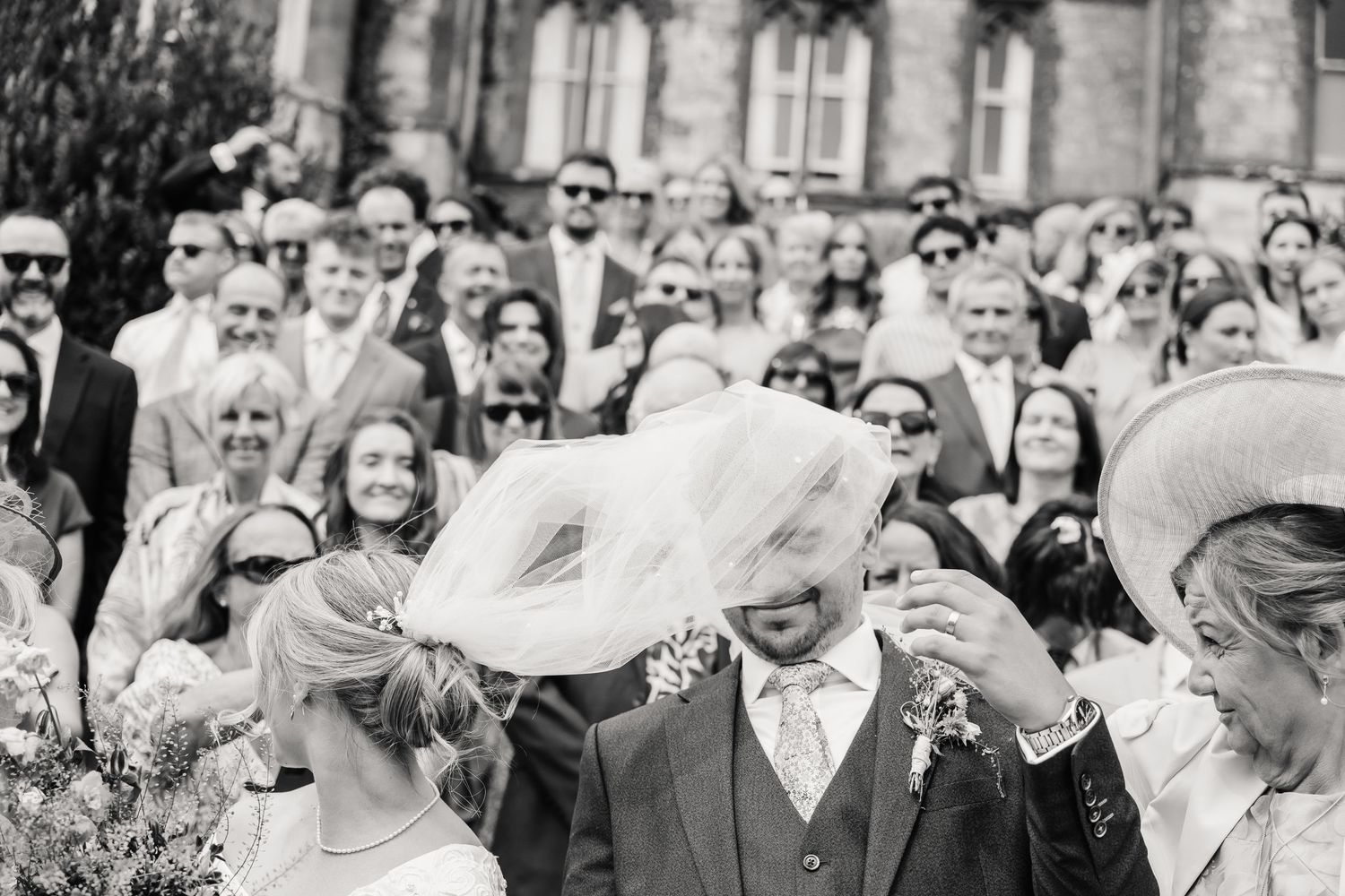 Black and white wedding photo of groom laughing as bride's veil blows over his face, guests watching behind them.