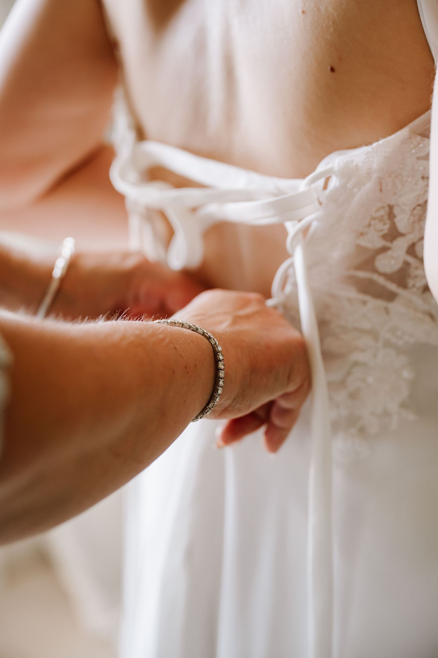 Helper tying lace-back wedding dress bow with bracelet-adorned hands at Pennsylvania Castle, Portland Dorset