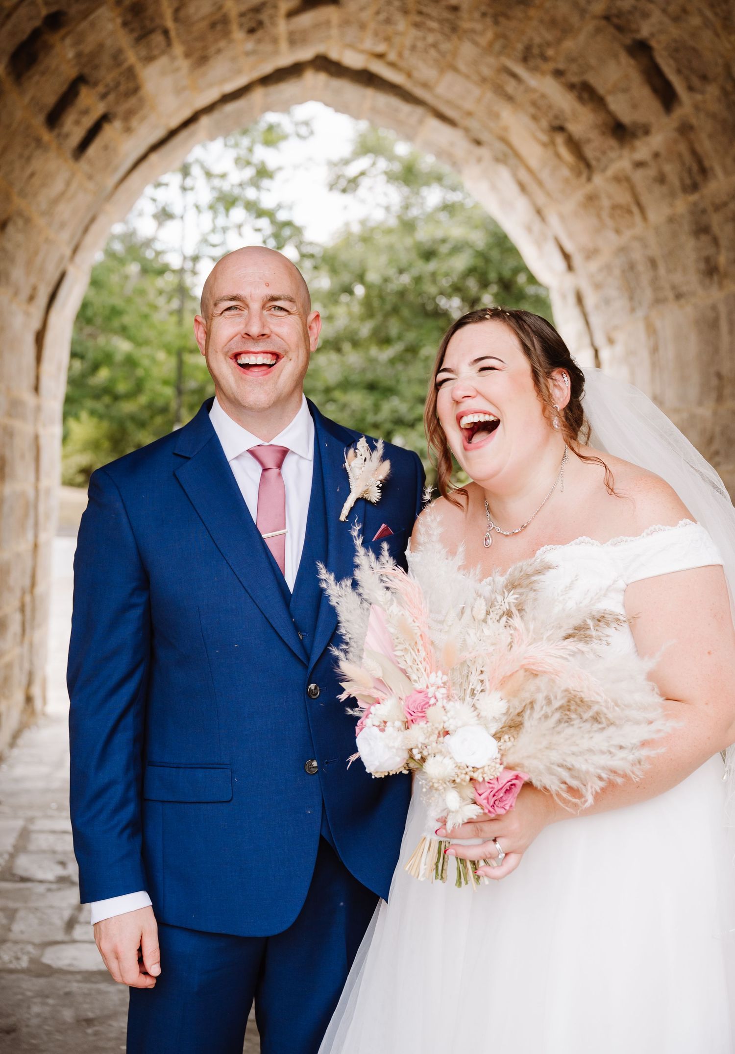 Laughing bride and groom pose under stone arch, bride holding pampas grass bouquet, groom in navy suit with pink tie.
