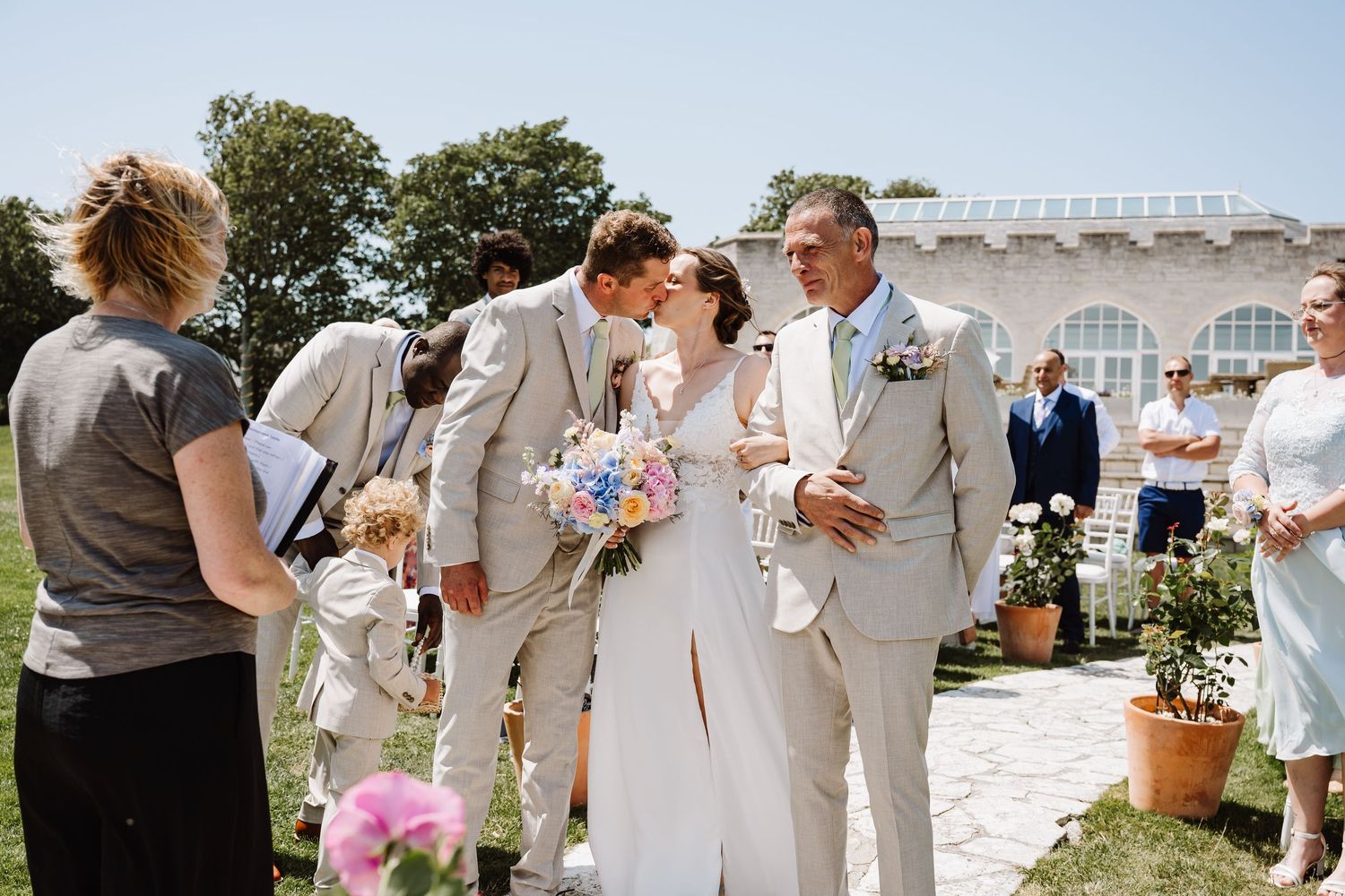 Newlyweds sharing a first kiss outdoors while guests celebrate in the background at Pennsylvania Castle, Portland Dorset