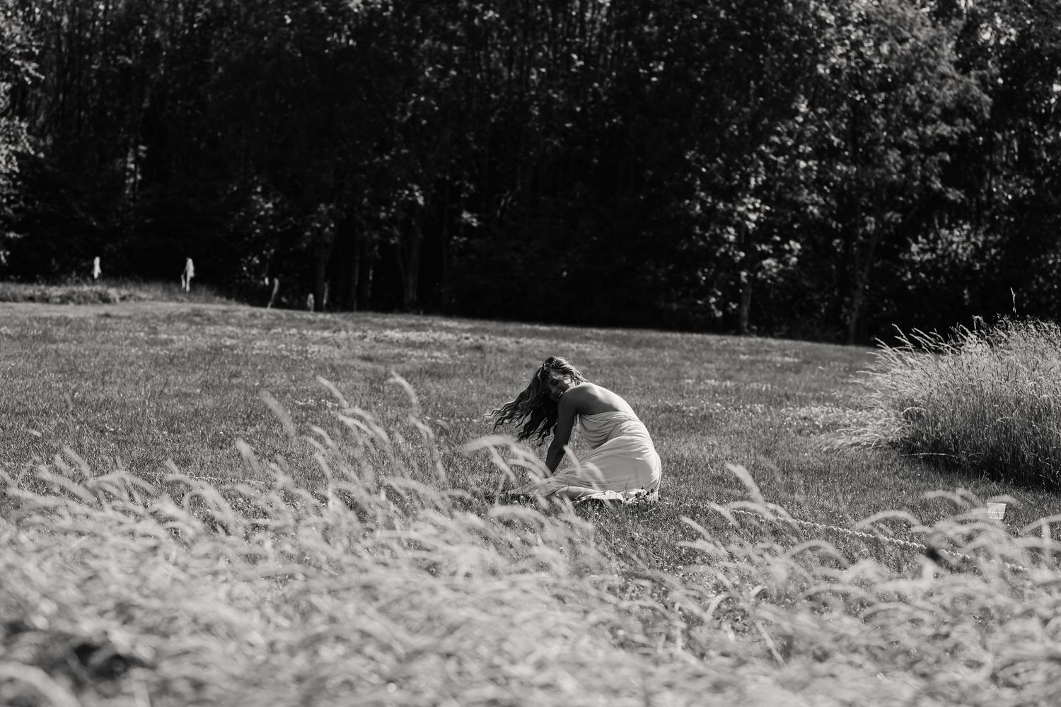 Black and white photo of a woman crouching in a grassy field with trees in the background.