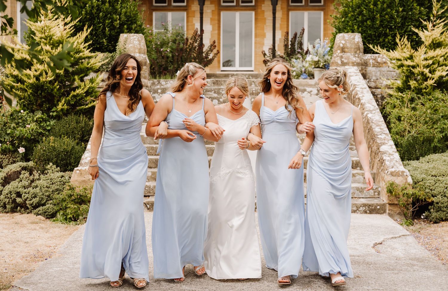 Bride and four bridesmaids in light blue gowns laughing together outside a stone venue.