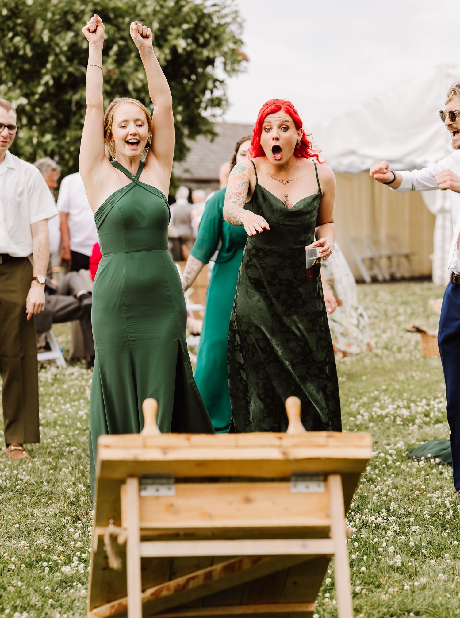 Two women in green dresses celebrate outdoors at a wedding lawn game, one raising arms in victory.
