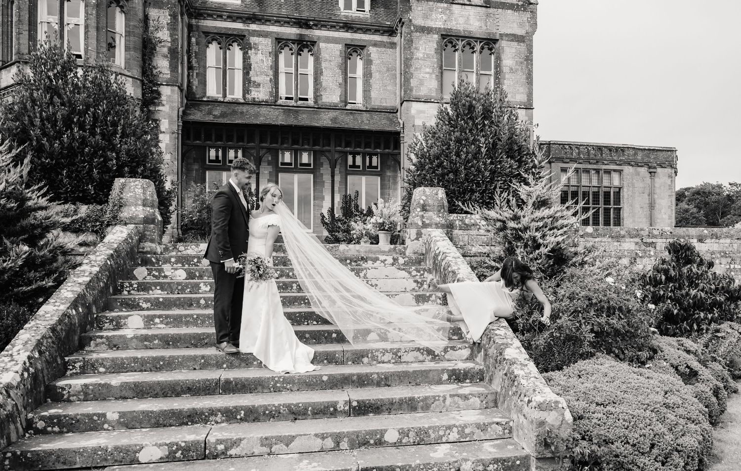 Black and white wedding photo of bride and groom on stone steps outside a grand historic manor house.