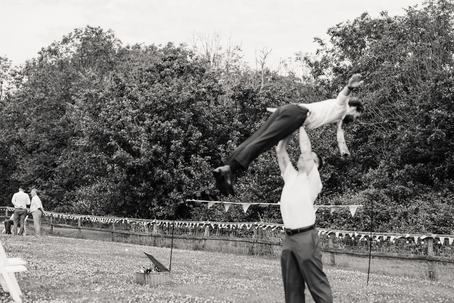 Black and white photo of a man lifting another person high in the air at an outdoor wedding.