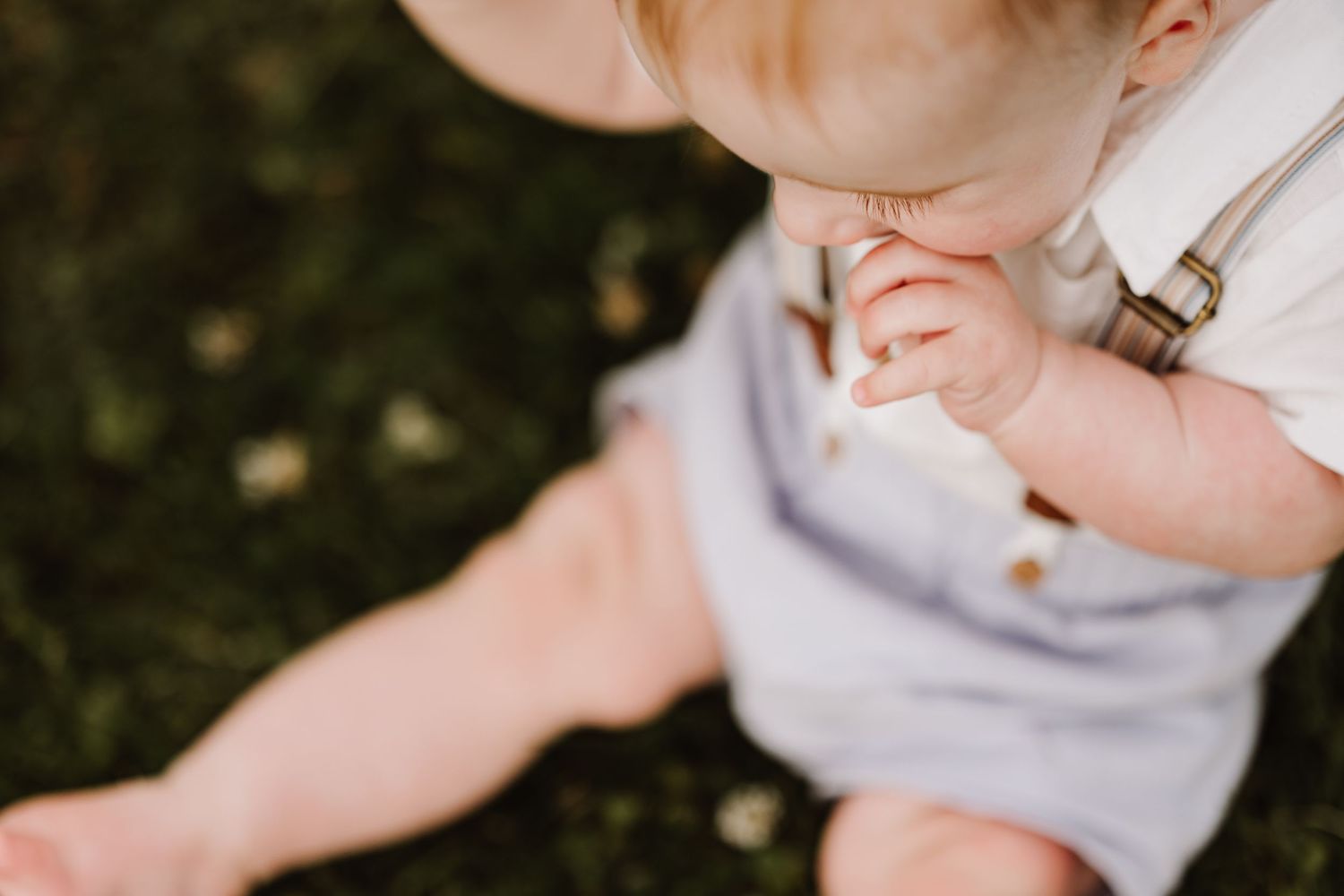 Adorable baby sitting on grass in light blue overalls, chewing on a snack outdoors.