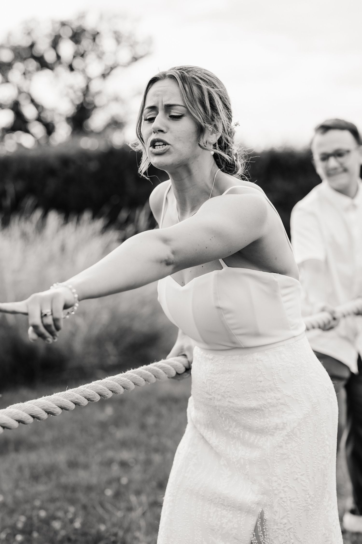 Bride in white dress straining during tug of war game at outdoor wedding reception, black and white photo.