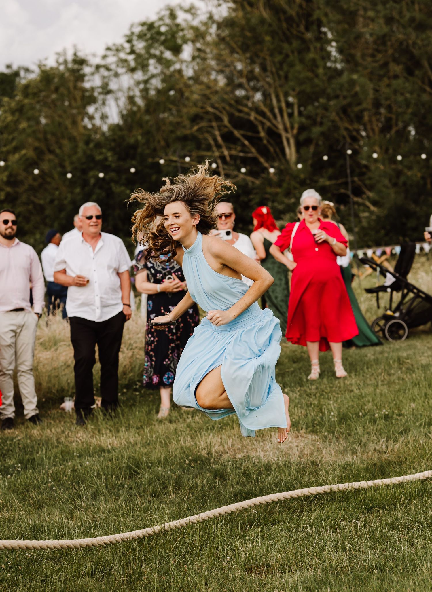 Woman in blue dress running during outdoor wedding lawn games while guests watch and cheer.