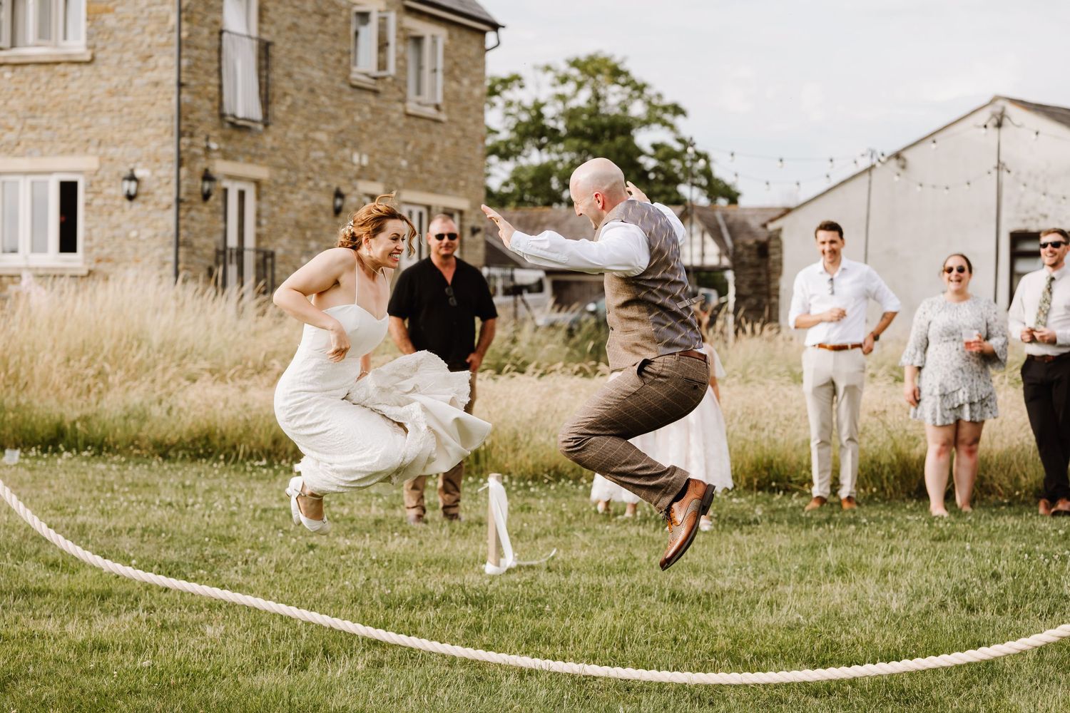 Bride and groom jumping rope together at outdoor wedding reception on green lawn with guests watching.