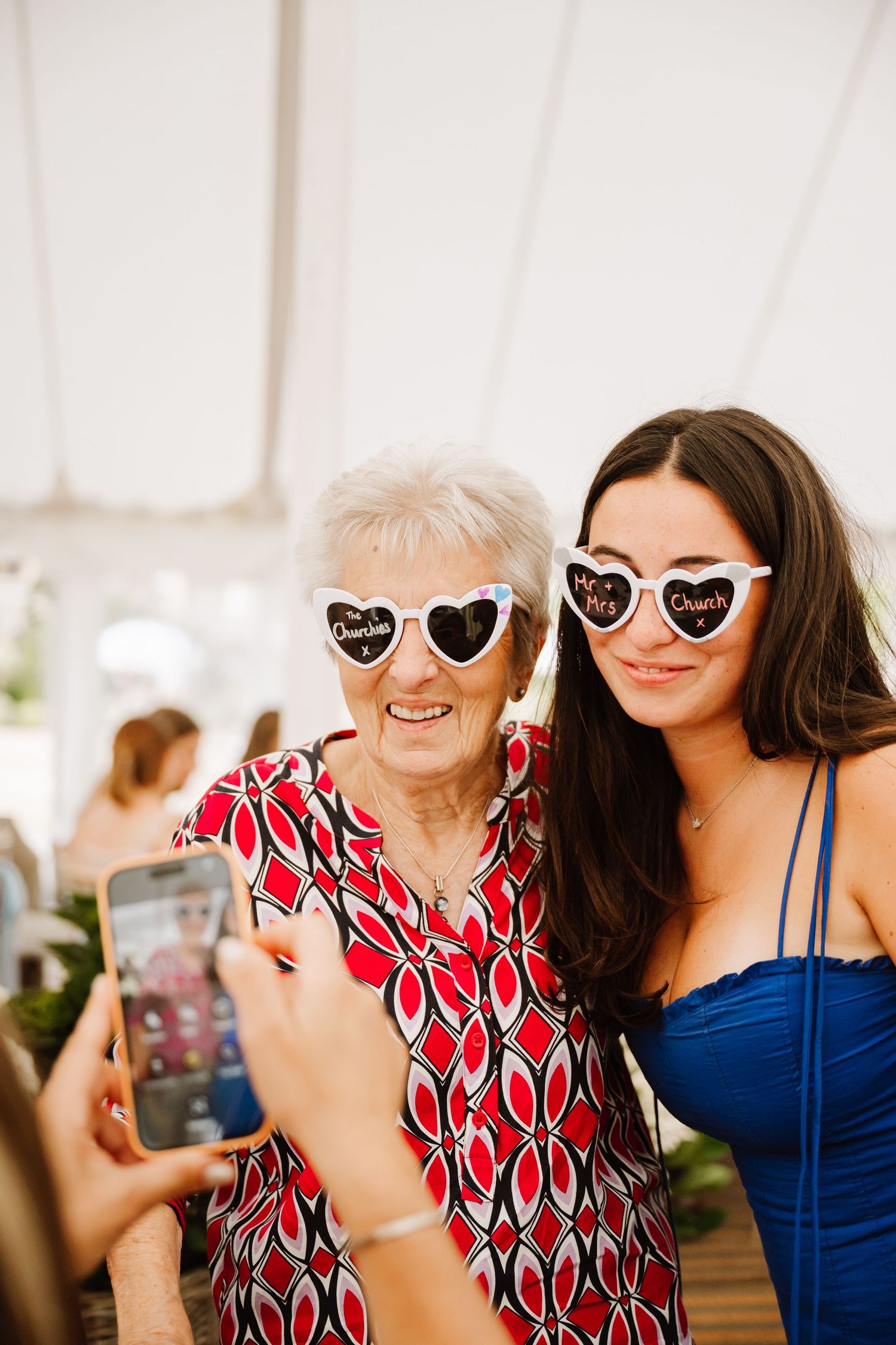 Elderly woman and young brunette pose for selfie wearing heart-shaped sunglasses at a sunny outdoor event.