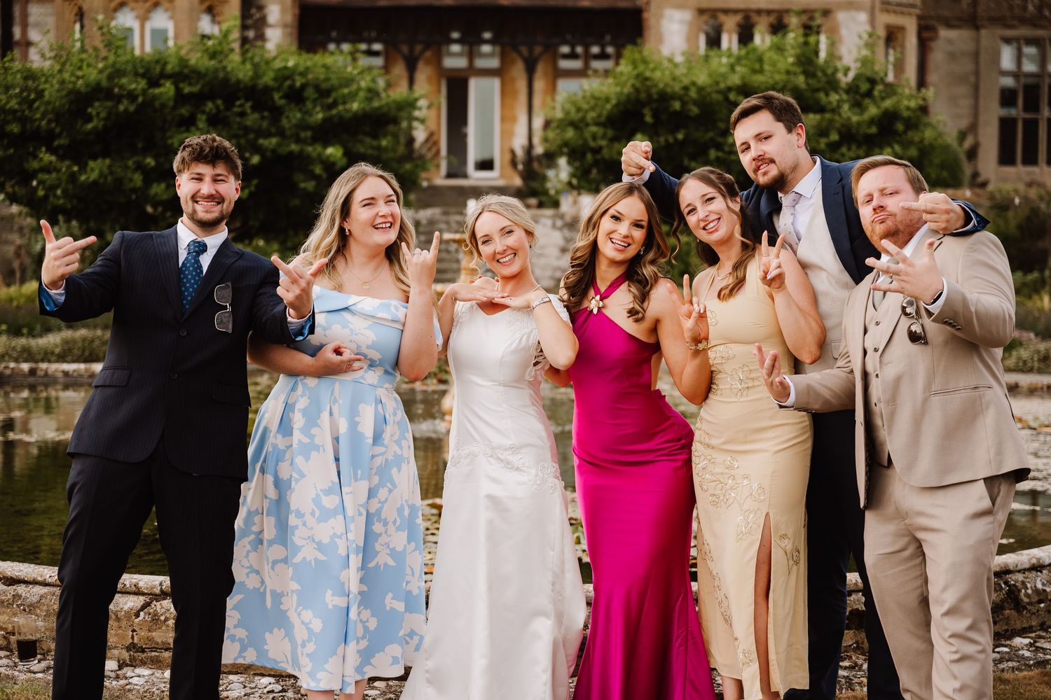 Wedding party of seven posing joyfully outdoors, bride in white gown with bridesmaids and groomsmen at elegant venue.