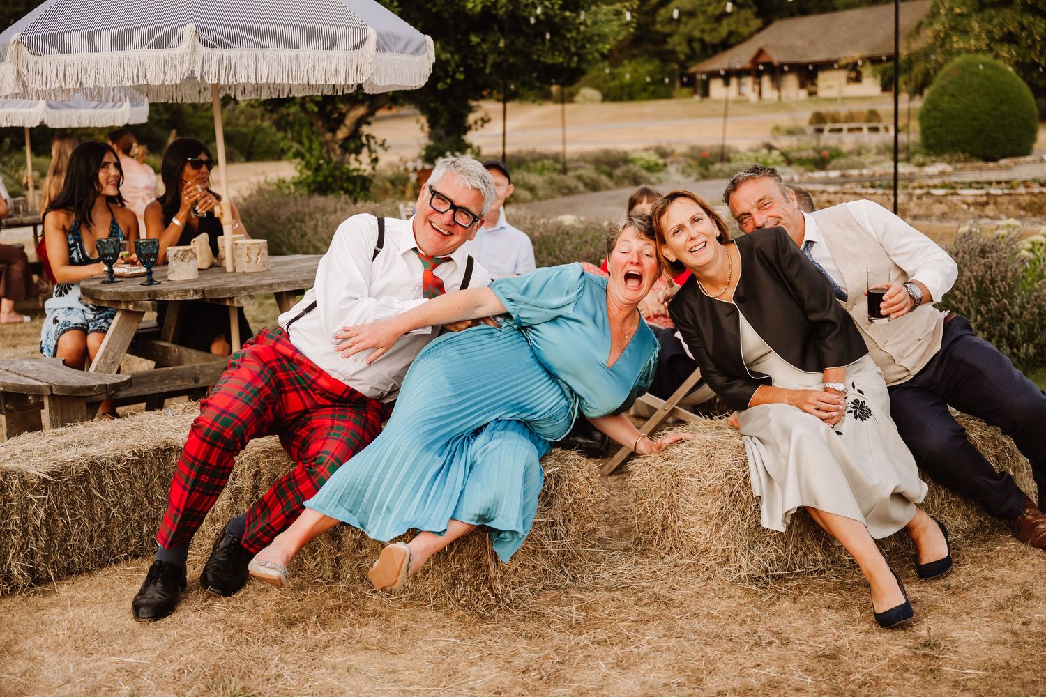 Group of friends laughing and relaxing on hay bales at an outdoor wedding celebration in a rustic garden setting.
