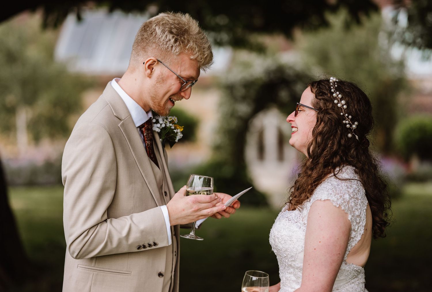 Bride and groom laughing together outdoors, holding champagne glasses, bride wearing floral crown, groom in tan suit.