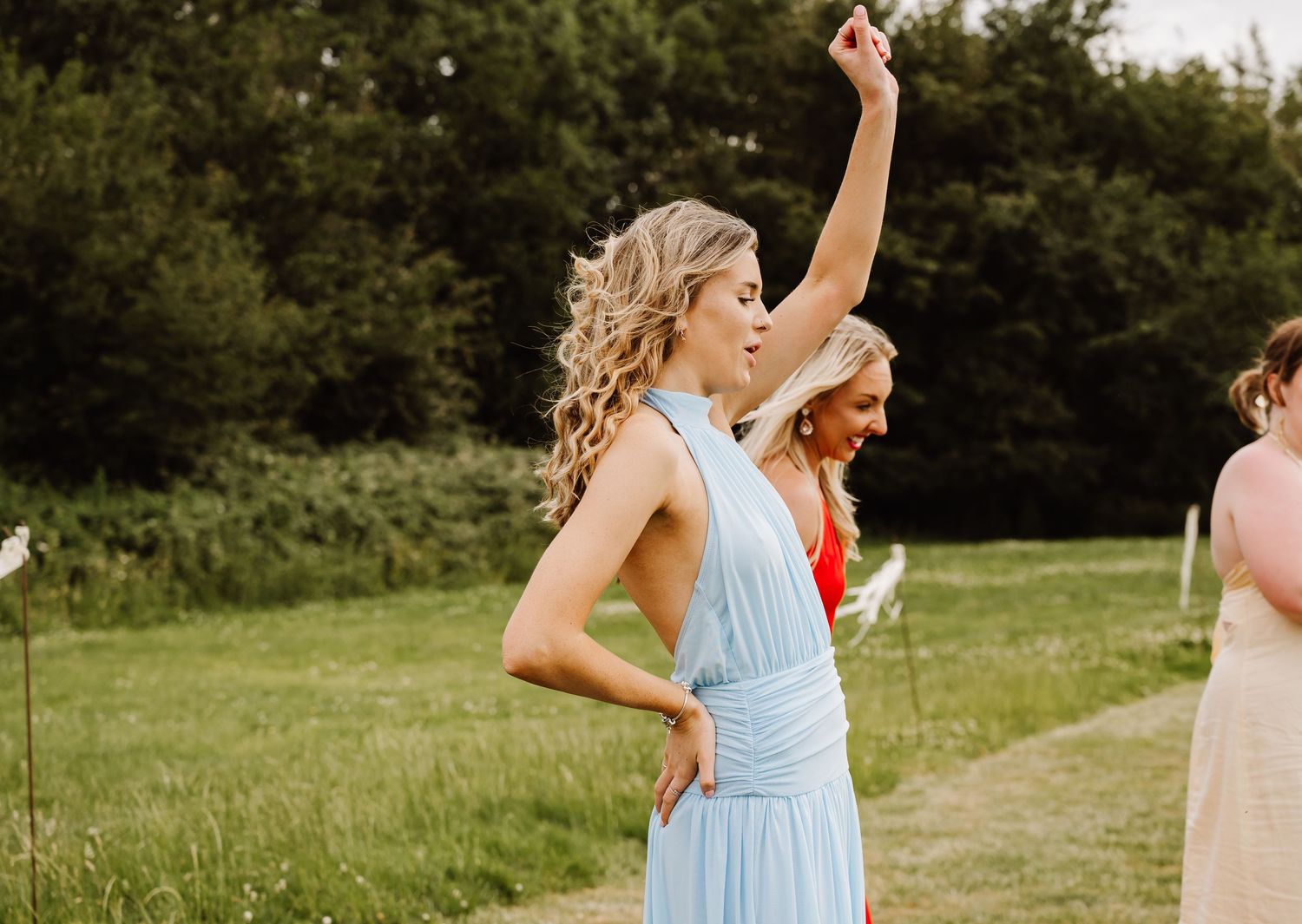 Bridesmaid in light blue dress dancing outdoors at a wedding with greenery in background.