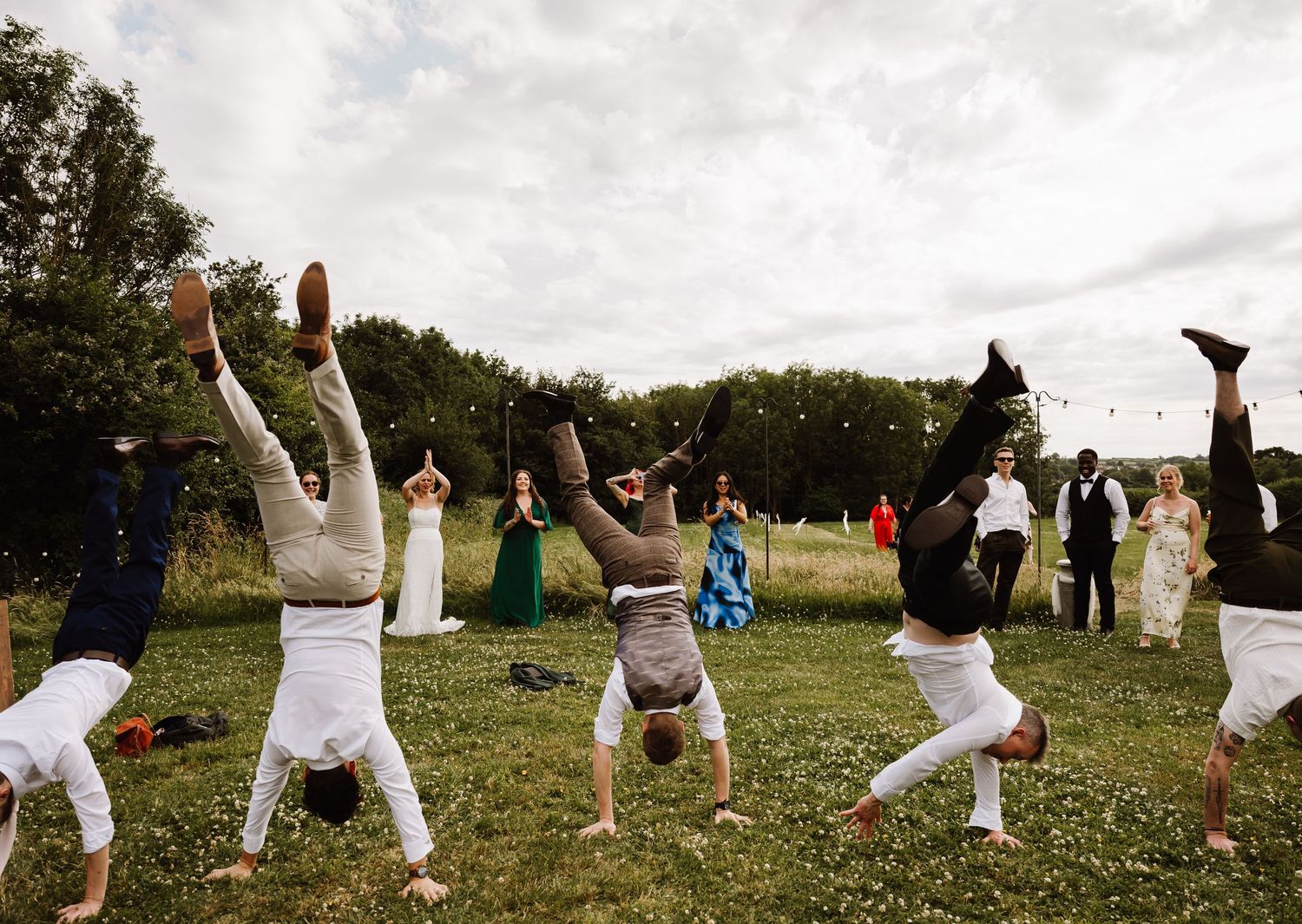 Wedding guests performing handstands on green grass at an outdoor wedding reception celebration.